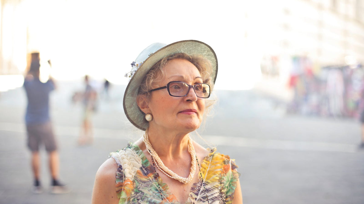 Femme de plus de 60 ans portant lunettes, rouge à lèvres rose, chapeau et collier perlé